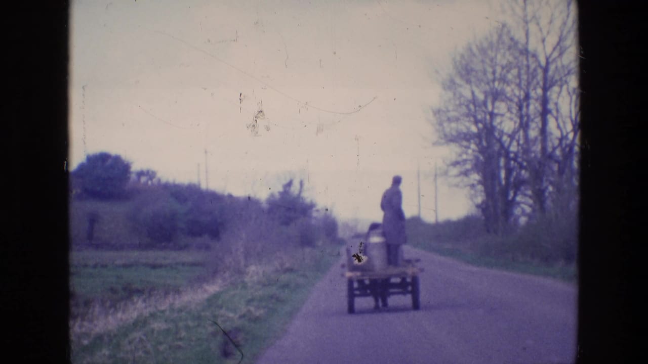 1969 Ireland home-movie frame showing a man standing on a small cart as it travels down a quiet rural paved road.