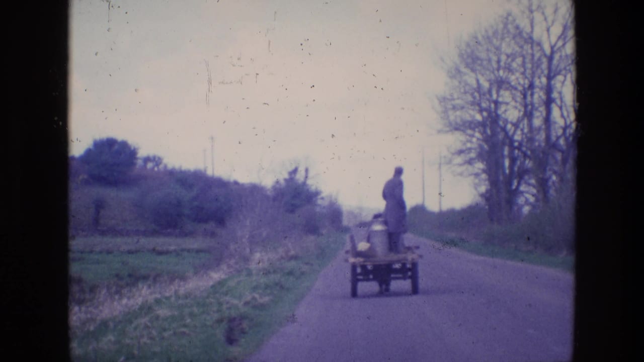 Small cart farther down a rural road with a standing man, grassy fields, hedges, and bare trees nearby.
