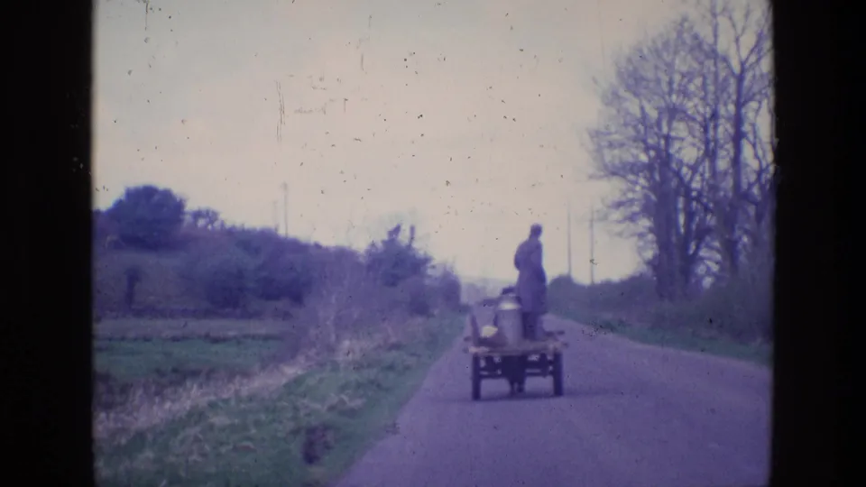 The cart and standing man continue away from the camera on an Irish rural road, with fields and leafless trees in view.