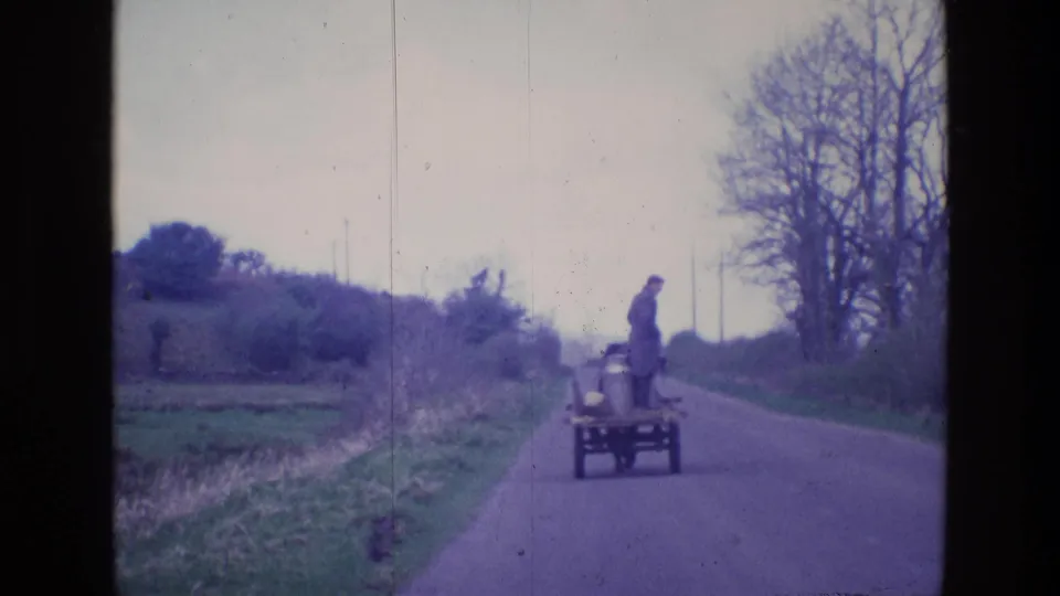 In this 8mm Ireland frame, a man in a long coat rides standing on a small cart along a quiet rural road.