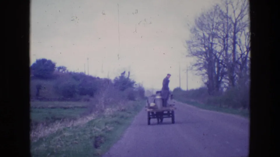 The man and small cart appear farther away on the Irish country road, with hedges, grass, and leafless trees lining the lane.