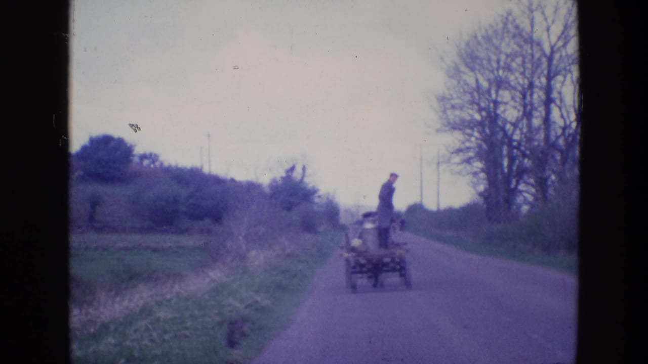 In 1969 Ireland footage, a uniformed man stands on a small utility cart moving down a narrow rural road lined with grass and bare trees.