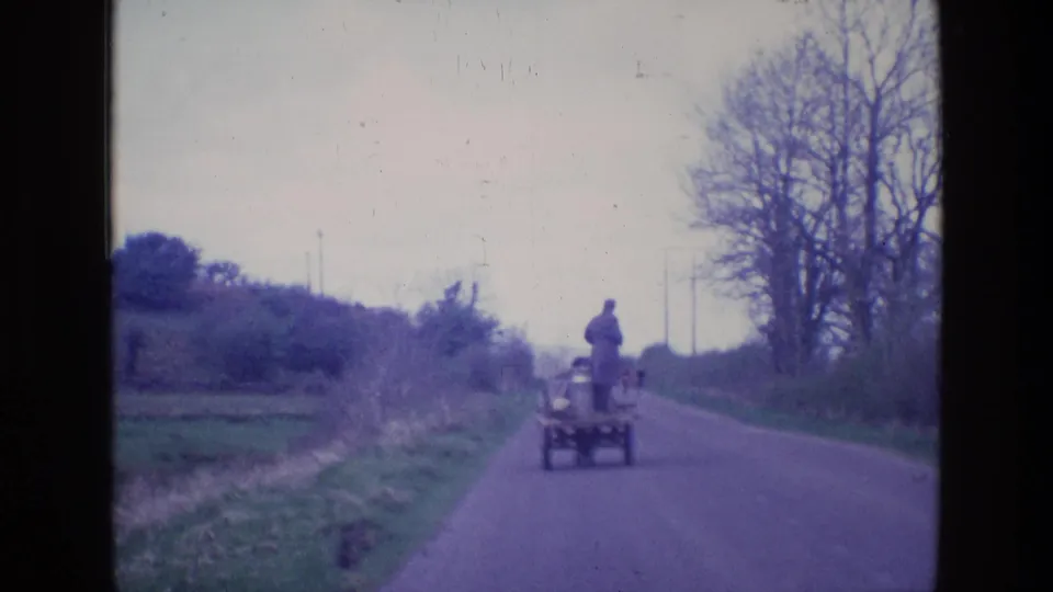 A uniformed man on a small utility cart travels farther from the camera on a narrow road bordered by grass, hedges, and bare trees.