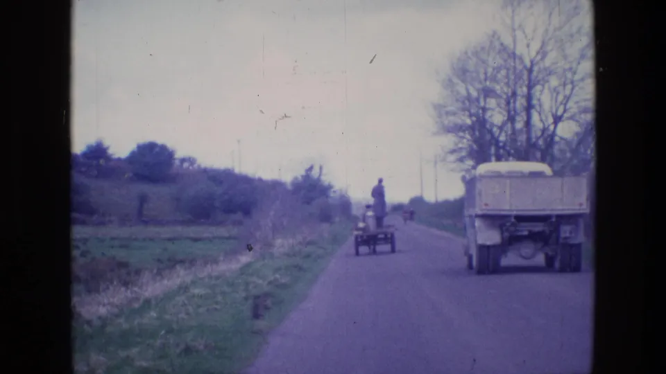 A larger truck enters the road foreground while the small utility cart with the standing man continues ahead in the distance.