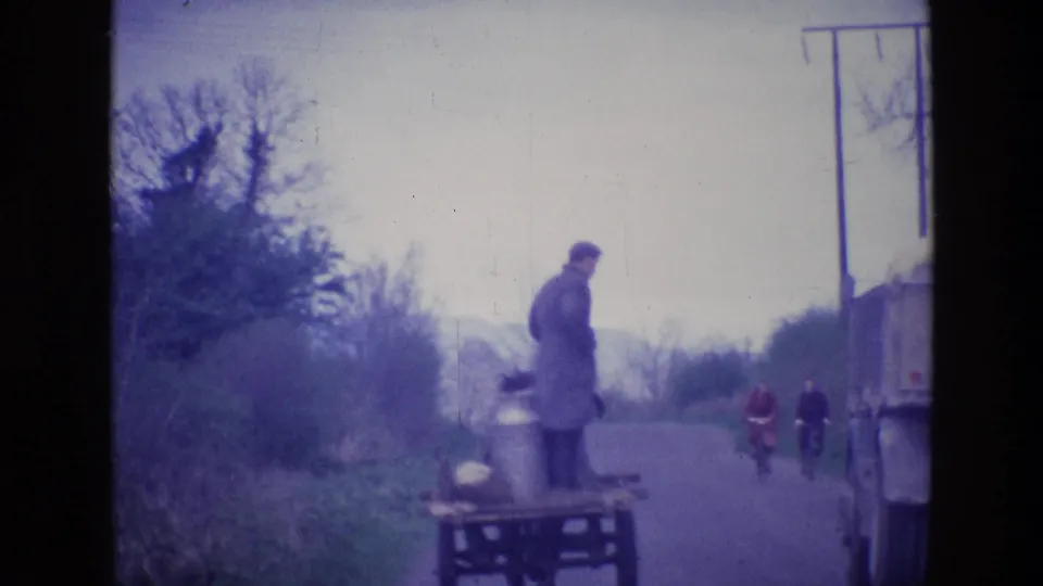 1969 Ireland rural road scene with a man standing on a cart, cyclists in the distance, and a truck entering the frame.