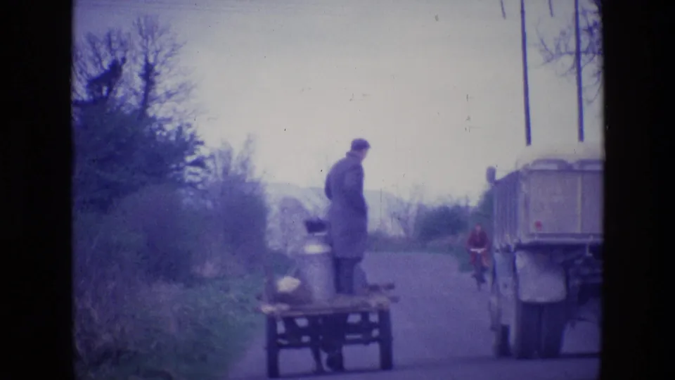 1969 Ireland frame showing the standing man on the cart closer to a large truck, with a cyclist farther down the road.