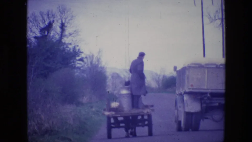 1969 Ireland home-movie frame with the man standing on the cart next to a truck on a narrow rural road.