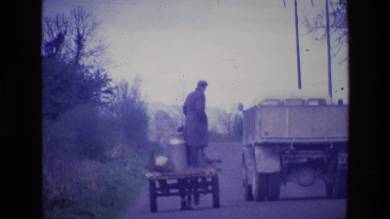 In rural Ireland in 1969, a man in a long coat stands by a horse-drawn cart loaded with milk cans while a truck passes.