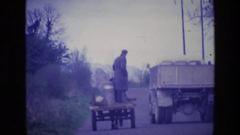 In rural Ireland in 1969, a man in a long coat stands by a horse-drawn cart loaded with milk cans while a truck passes.