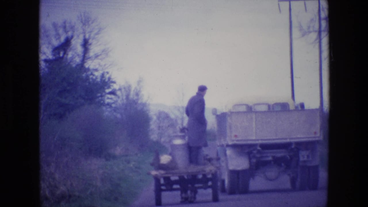 Truck passing a roadside cart while a man stands near milk cans on a narrow country road.