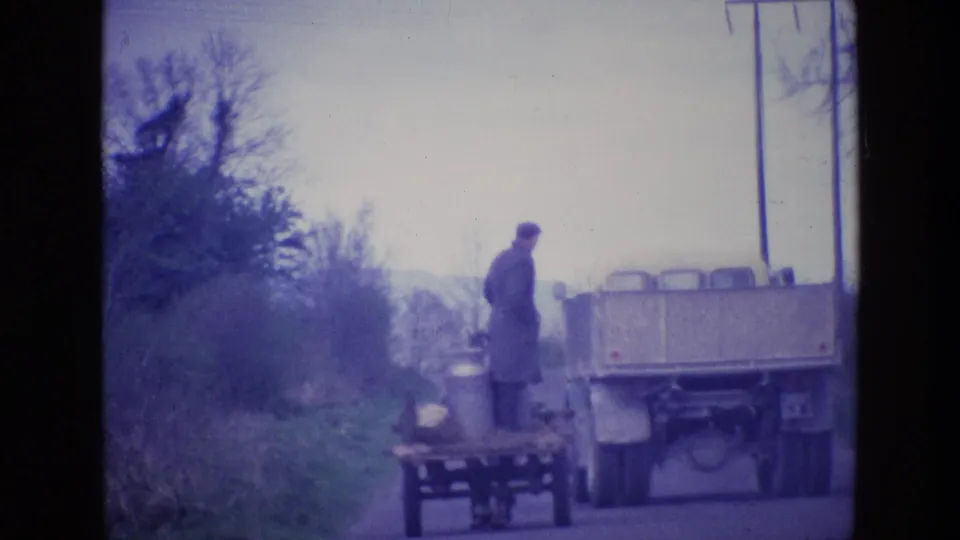 A 1969 rural Ireland roadside view shows a man beside a small cart with milk cans as a truck passes close by.