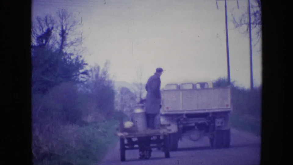 The man stands beside the horse-drawn milk cart while the rear of the truck fills the road in this 1969 Ireland home-movie frame.