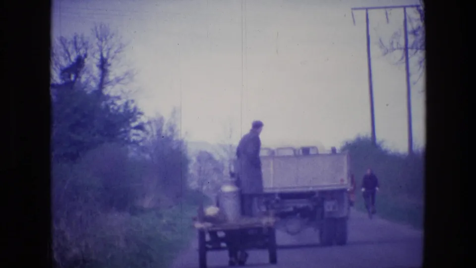 A rural Ireland road scene from 1969 shows the man, cart, milk cans, passing truck, and a distant bicyclist down the lane.