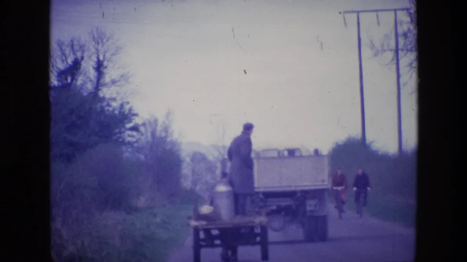 1969 Ireland 8mm frame showing a man on a wooden cart near a truck, with two cyclists behind on a narrow rural road.