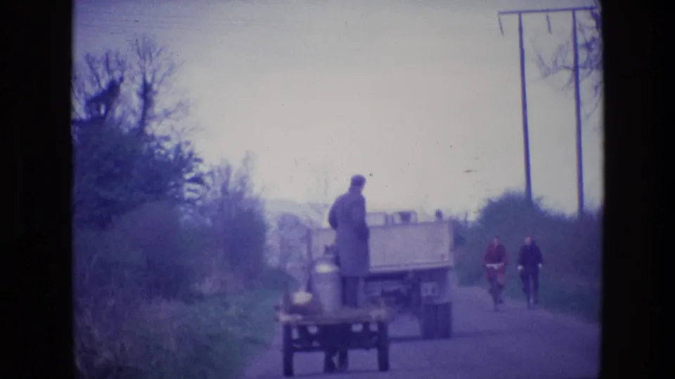 1969 Ireland 8mm frame with a truck and cart on a country road, two cyclists behind, and utility poles beside hedgerows.