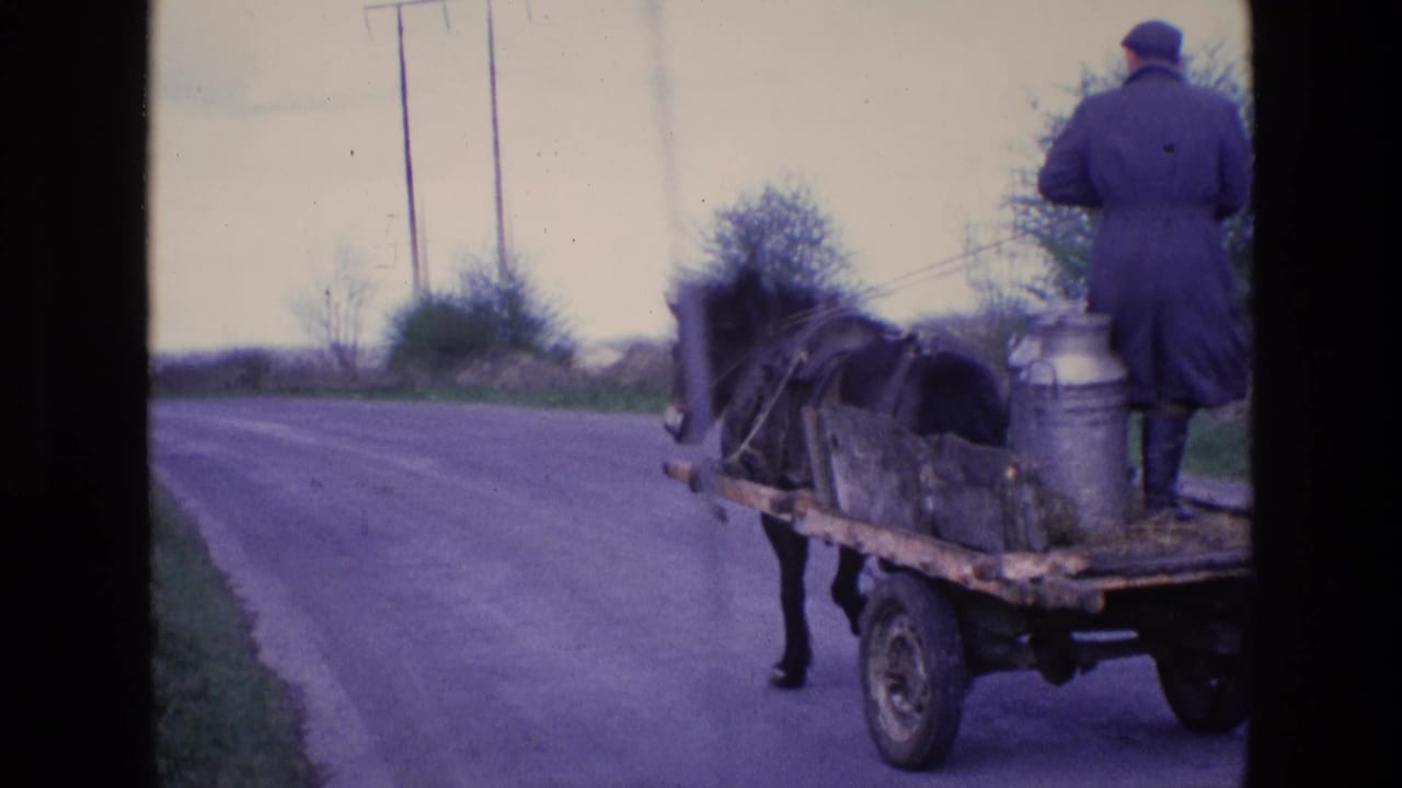 1969 Ireland home-movie frame showing a milkman from behind on a horse-drawn cart carrying metal milk cans along a countryside road.