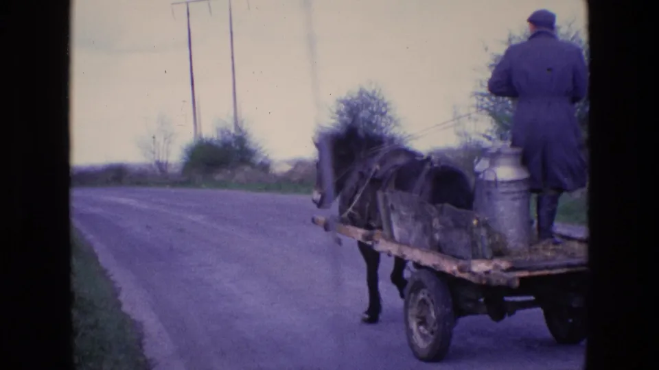 1969 Ireland home-movie frame showing a milkman from behind on a horse-drawn cart carrying metal milk cans along a countryside road.