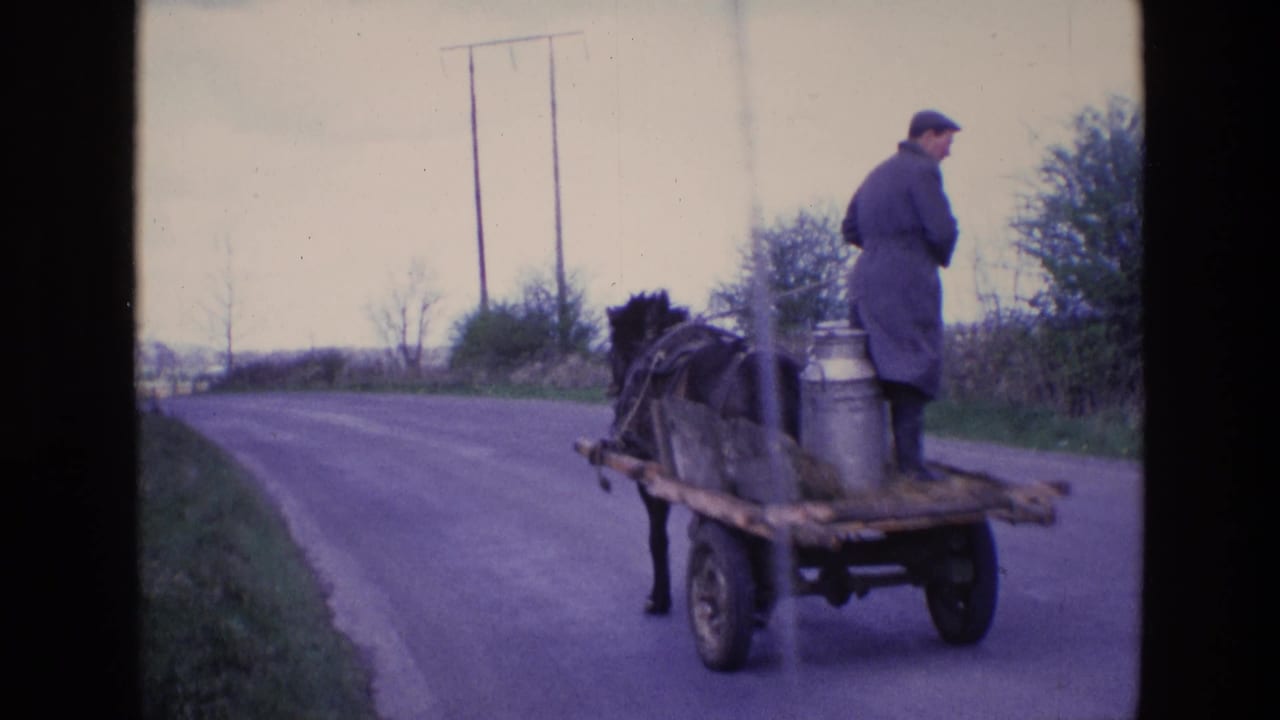 1969 Ireland frame of a milkman on a wooden horse cart with metal churns, viewed from behind on a rural paved road.