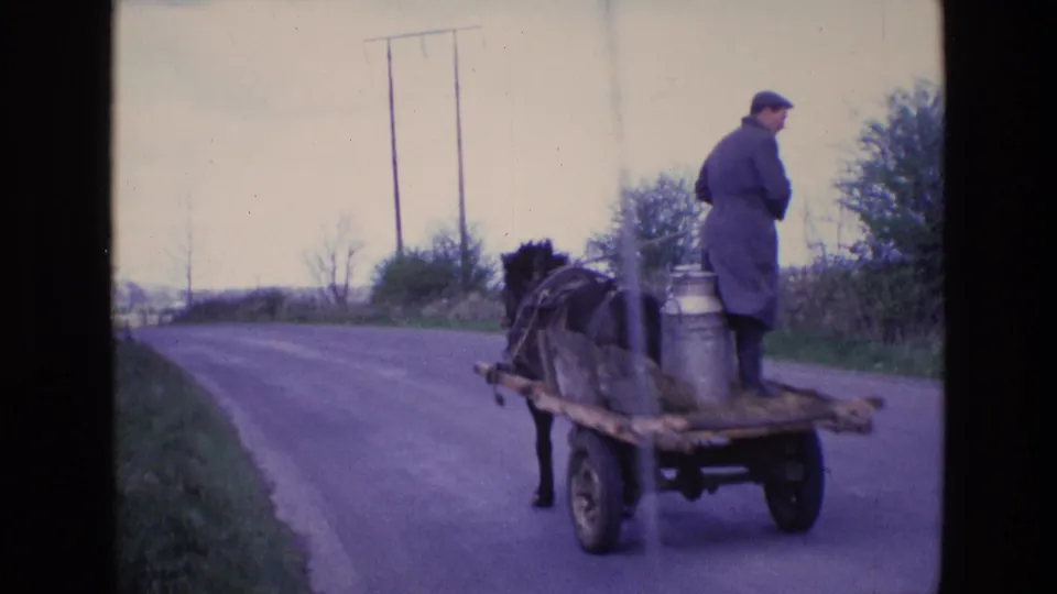 1969 Ireland frame of a milkman on a wooden horse cart with metal churns, viewed from behind on a rural paved road.