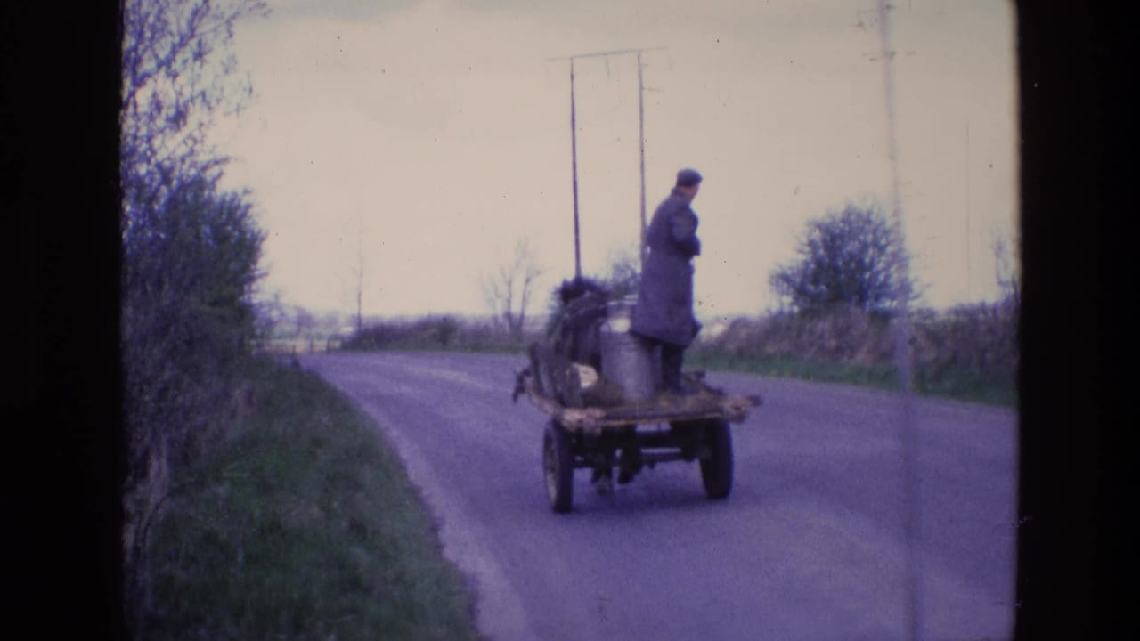 In 1969 Ireland, a man in a long coat and cap rides standing on a horse-drawn wagon with a metal milk churn along a hedged rural road.