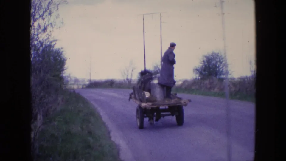 In 1969 Ireland, a man in a long coat and cap rides standing on a horse-drawn wagon with a metal milk churn along a hedged rural road.
