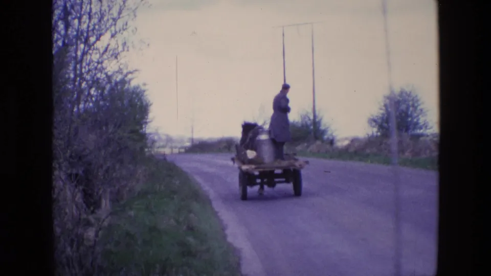 The horse-drawn wagon and standing rider continue away on an Irish country lane, framed by hedgerows, poles, and muted 8mm color.