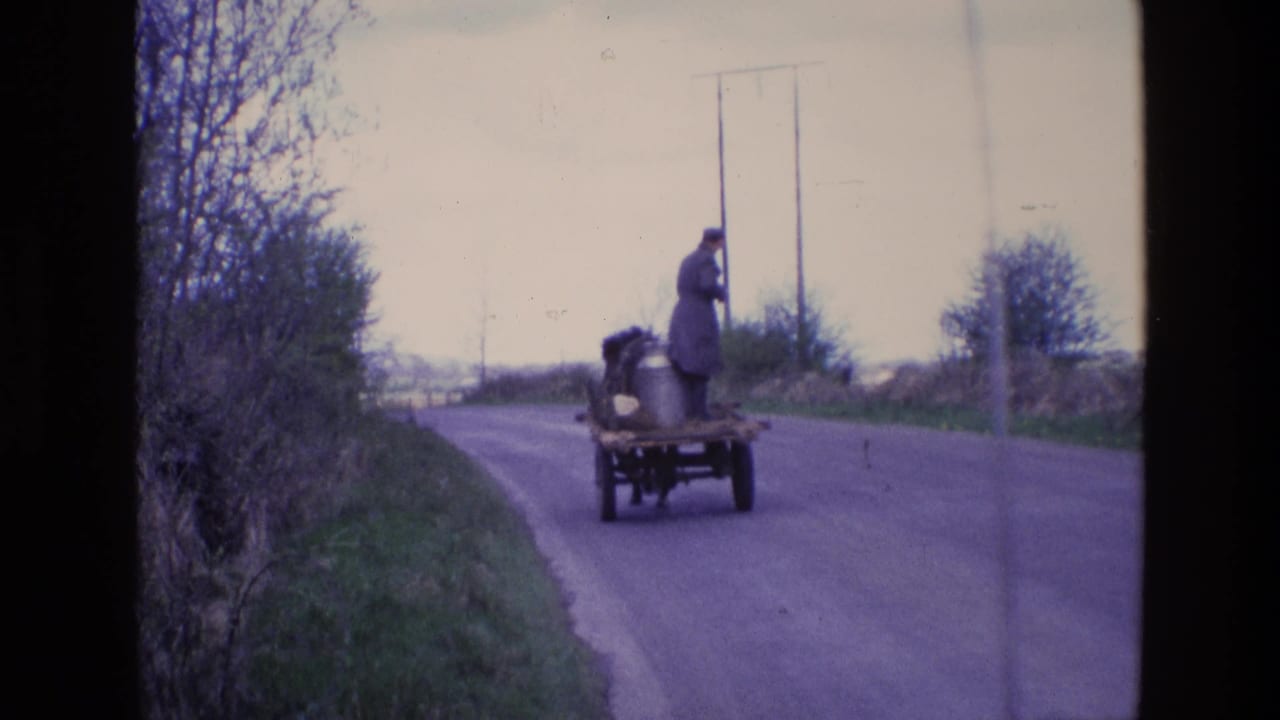 1969 Ireland home-movie frame showing a man standing on a horse-drawn cart with metal milk cans on a quiet rural road.