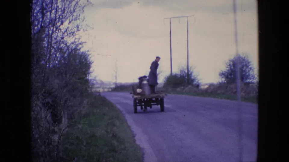 Late-1960s Irish rural scene with a horse-drawn milk cart receding along a hedge-lined road under a pale sky.