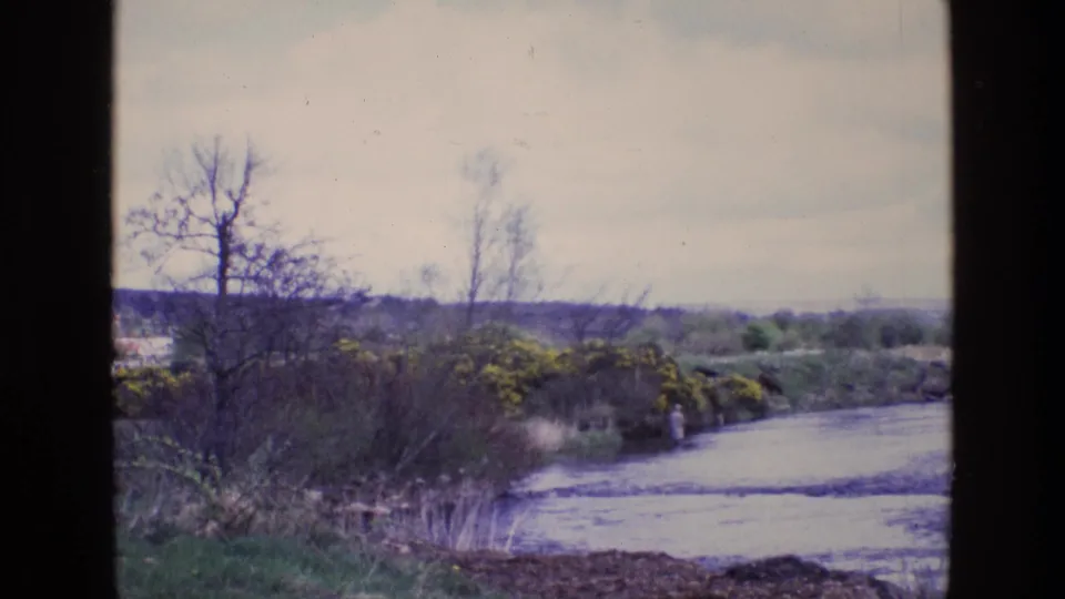 1969 Ireland 8mm frame showing a riverbank landscape with bare trees and a distant person near the water's edge.