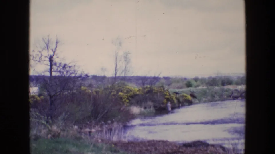 1969 Ireland 8mm frame of a lone distant person at a rural riverbank framed by bare trees, reeds, and faded sky.