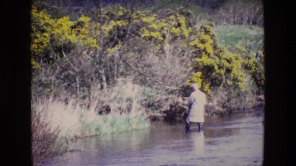 The 8mm frame shows a person in pale outerwear moving along the river edge in rural Ireland, with reeds and yellow shrubs behind.
