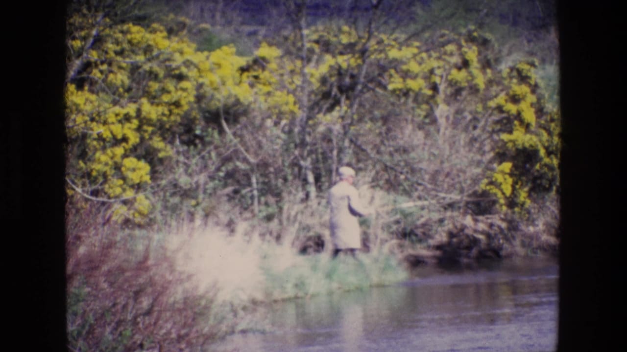 Man in a light coat fly fishing beside a river with yellow bushes behind him.