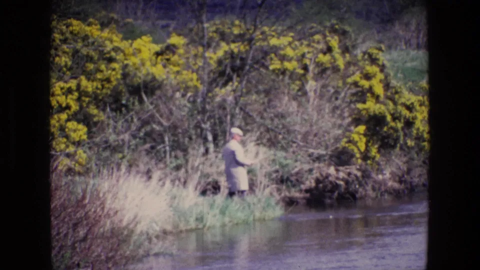 The fisherman remains by the Irish riverbank, framed by spring grasses, yellow gorse, and hazy 8mm color.