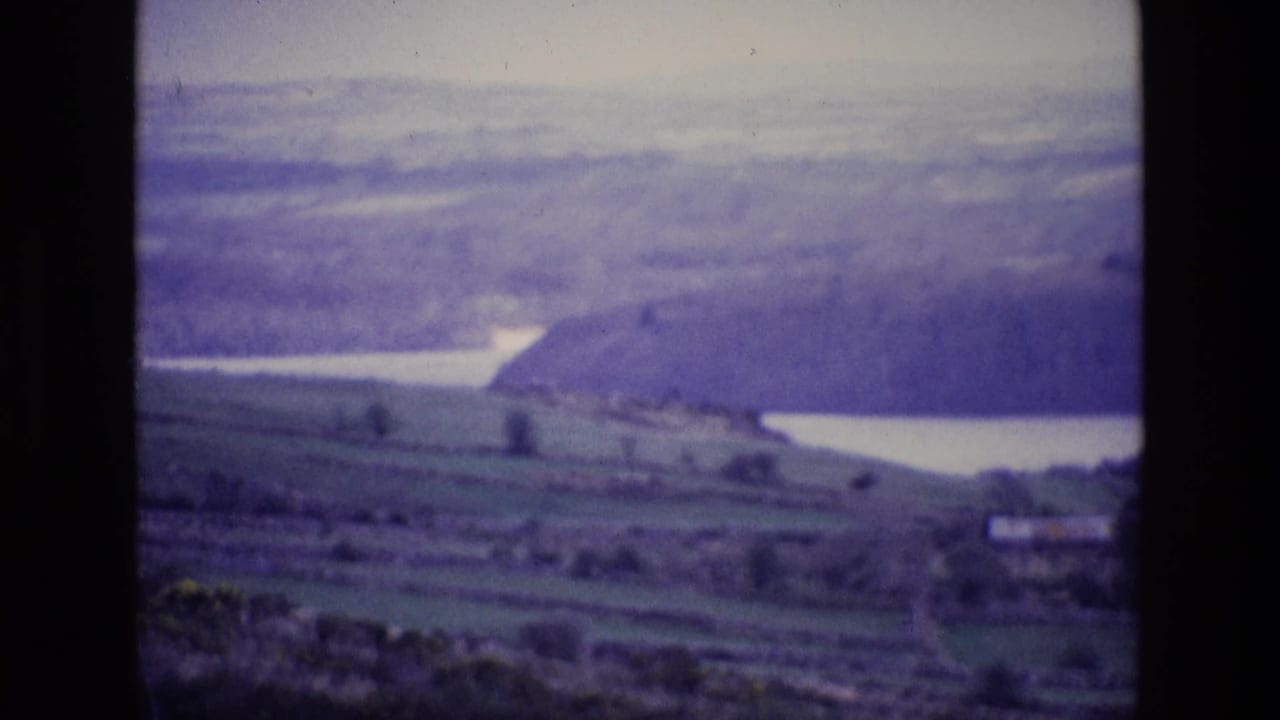 1969 8mm view of Irish countryside with green fields, a pale lake, wooded slope, and hazy rolling hills.