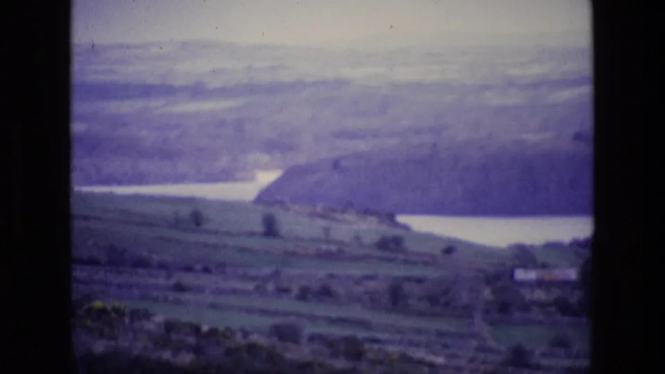 1969 8mm view of Irish countryside with green fields, a pale lake, wooded slope, and hazy rolling hills.