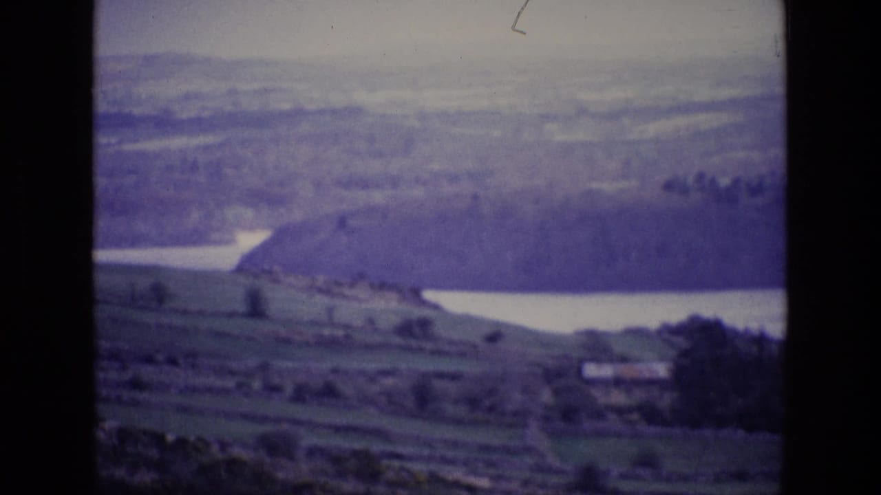 1969 Irish countryside frame showing divided fields, a calm lake, wooded hillside, distant hills, and a small valley building.
