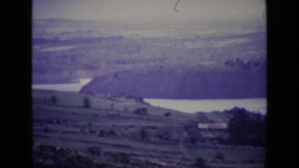 1969 Irish countryside frame showing divided fields, a calm lake, wooded hillside, distant hills, and a small valley building.