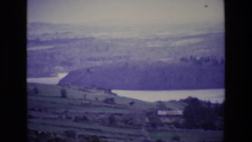 1969 8mm frame of Irish rural fields sloping toward a pale lake, with wooded ground and misty hills beyond.