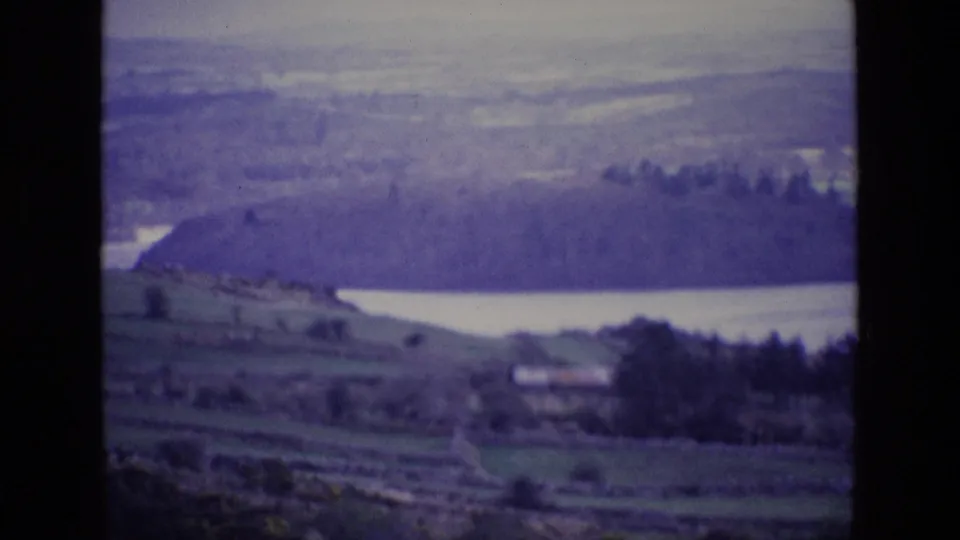 1969 Irish landscape frame with a lake, green fields, dark trees, a small building, and soft distant hills.