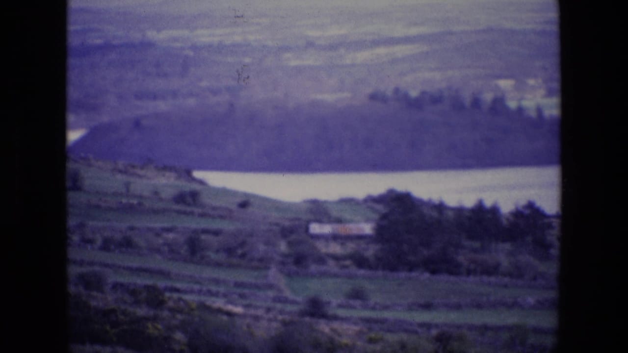 1969 Ireland home-movie frame showing a lake beyond terraced green fields, trees, and a small rural building.