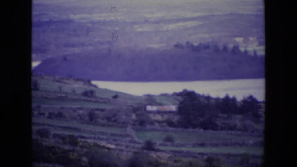 1969 Ireland home-movie frame showing a lake beyond terraced green fields, trees, and a small rural building.