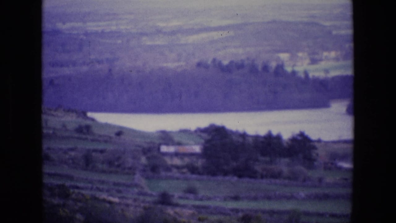 1969 Ireland countryside view with a pale lake, dark wooded far shore, fields, and a small building in the lower landscape.