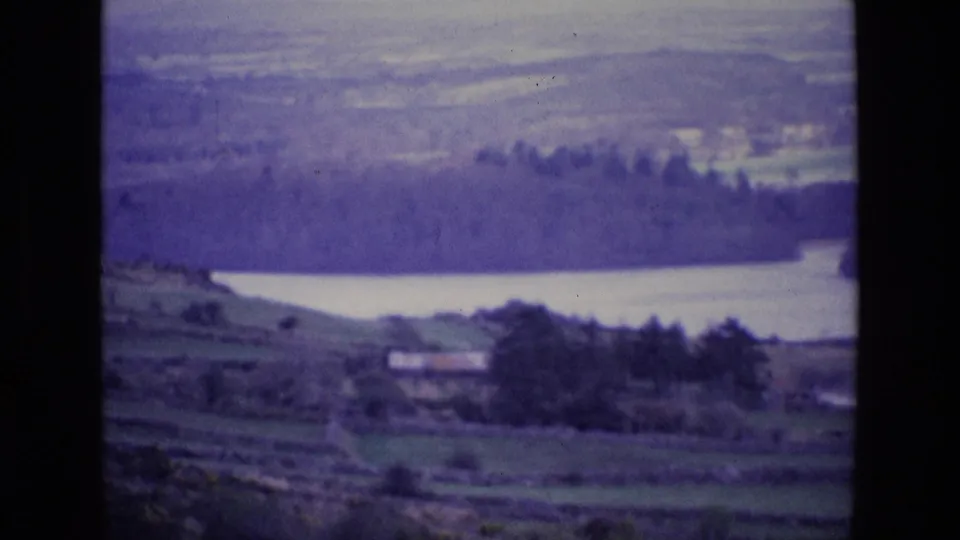 1969 Ireland countryside view with a pale lake, dark wooded far shore, fields, and a small building in the lower landscape.