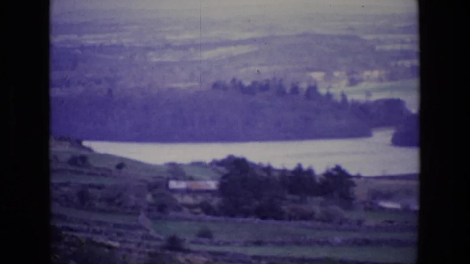 1969 Ireland frame showing layered pastures, a rural building, tree cover, and a lake backed by hazy hills.