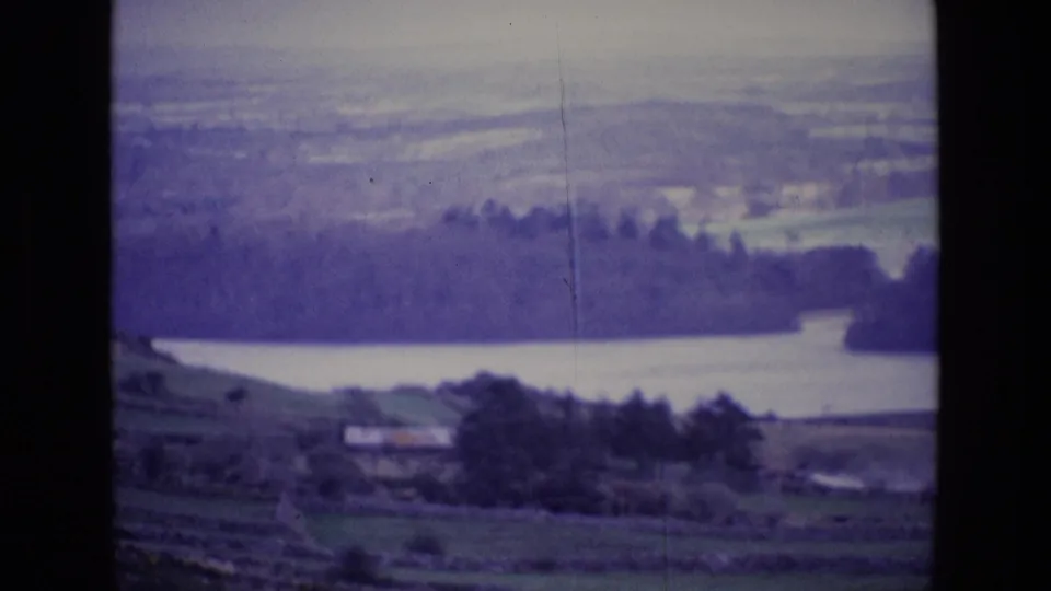 1969 Ireland home-movie frame with green fields and a small building below a lake, wooded shoreline, and muted distant hills.