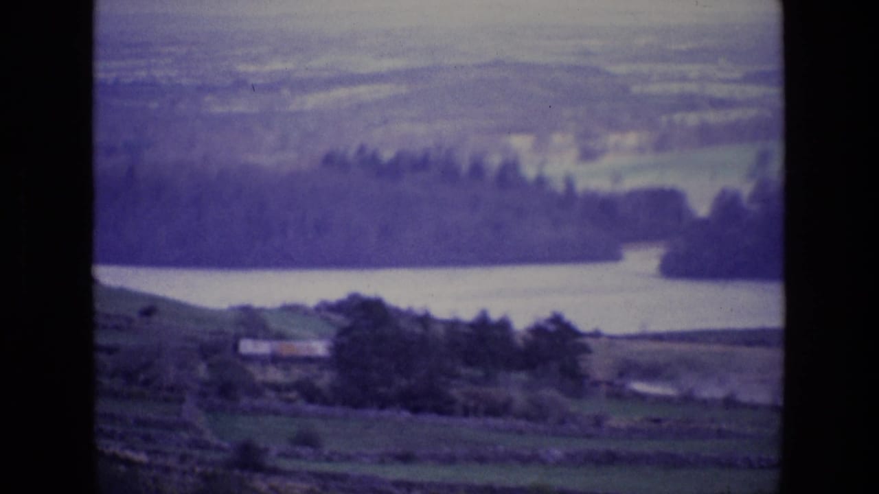 1969 Ireland 8mm frame showing a lake below rolling hills, with fields, stone walls, trees, and a small distant structure.