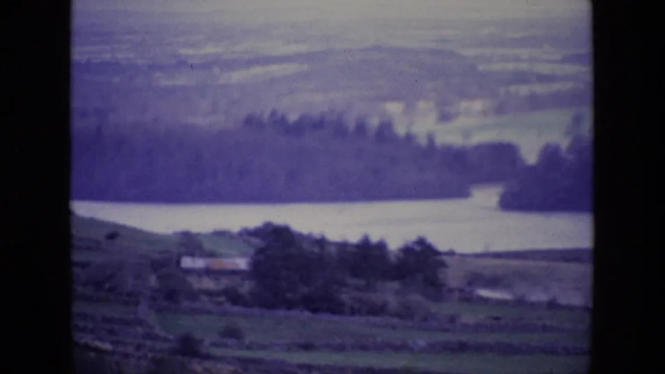 1969 Ireland 8mm frame showing a lake below rolling hills, with fields, stone walls, trees, and a small distant structure.