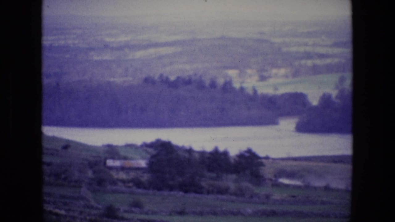 Vintage Ireland landscape frame with a broad lake, tree-lined shore, rolling fields, and hazy hills in the distance.