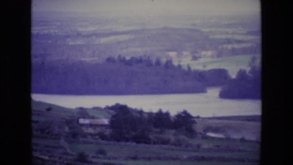 Vintage Ireland landscape frame with a broad lake, tree-lined shore, rolling fields, and hazy hills in the distance.