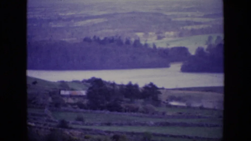1969 Ireland home-movie frame showing a quiet lake below wooded hills and rural fields divided by stone walls.
